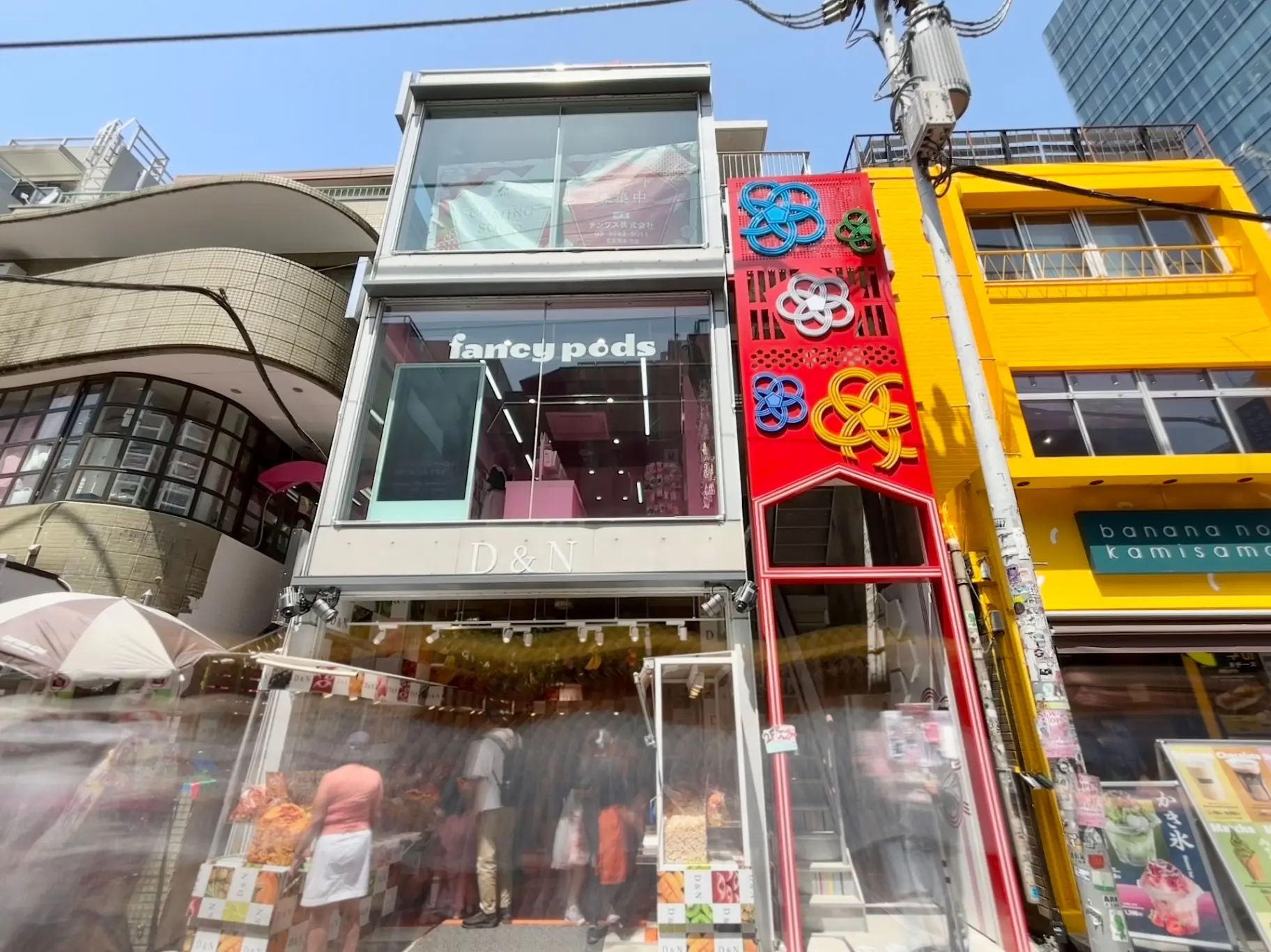 - おしゃれな店舗が並ぶ通りの風景、赤い装飾とガラス張りのファサードを持つD&N店舗が目立つ、賑やかな人々が行き交う様子