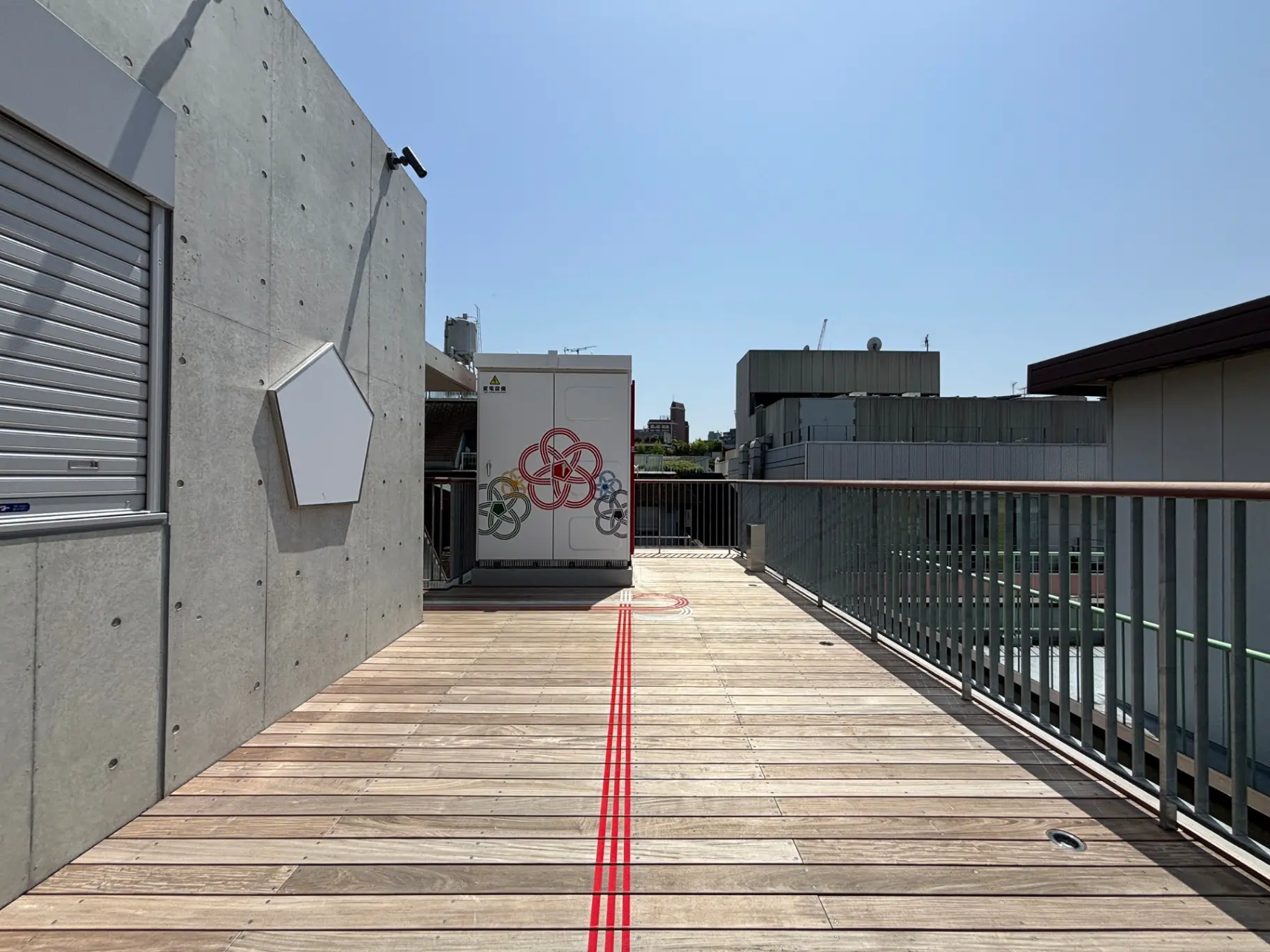 - 明るい青空の下、木製のデッキとコンクリート壁のある屋上スペース、六角形のアートが施された収納ボックスが配置された風景