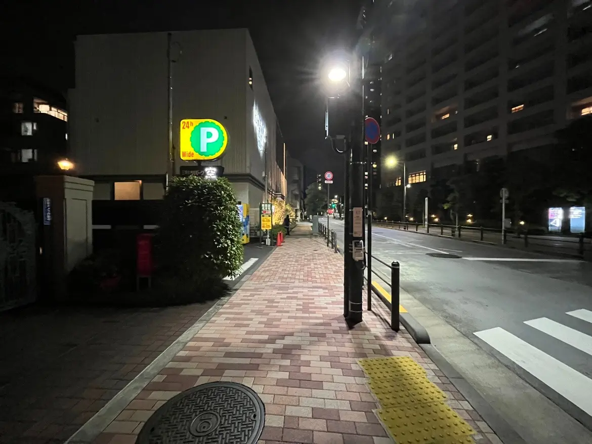 - 夜の静かな街並み、明るく照らされた駐車場の看板と舗装された歩道、周囲のビルが立ち並ぶ風景