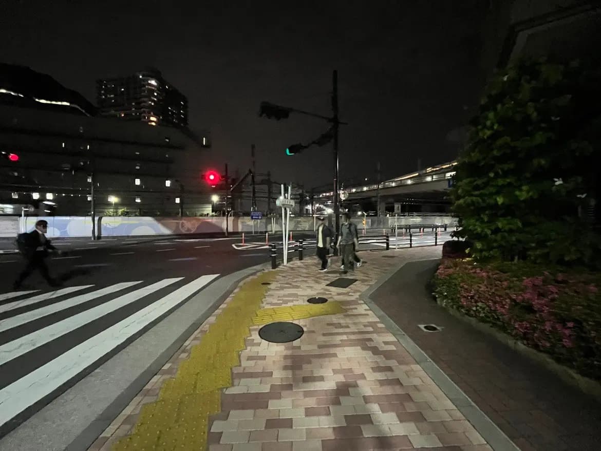 夜の街角、信号待ちの人々と道路、近くの建物と街灯が映える街の風景。