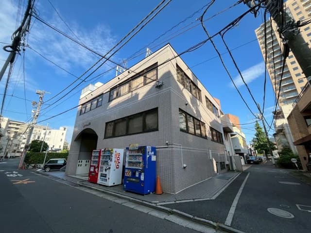 - 明るい青空の下、飲料自販機が並ぶ街角のビルと電柱、周囲に住宅が立ち並ぶ風景