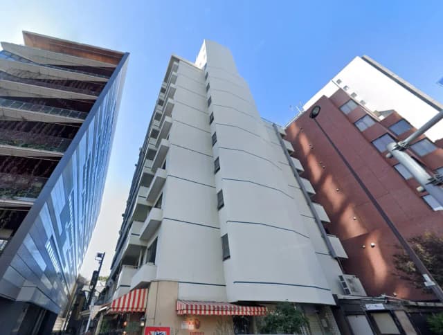 - 都市の高層ビル群の中に立つ白い外観のマンション、近くには赤レンガの建物と店舗が見える景観