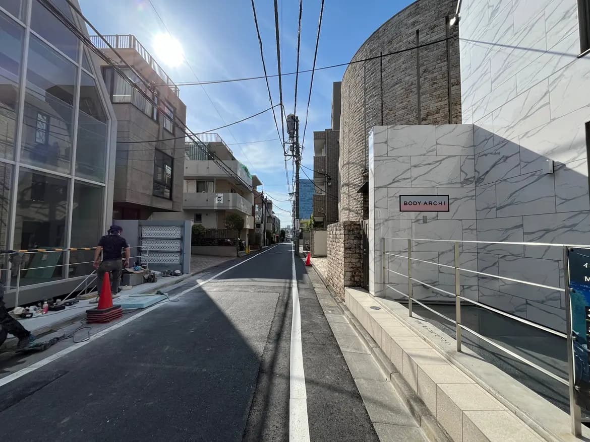 - 明るい日が差し込む舗装された通り、近代的な建物とBODY ARCHIの看板、工事作業者がいる風景