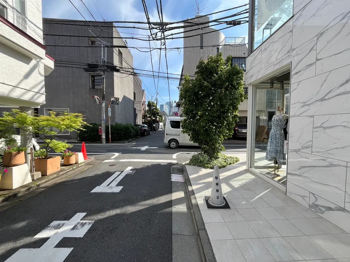 - 明るい青空の下、シンプルでおしゃれな商業ビルの角に立つショップ、対面には道路と緑の植栽がある風景