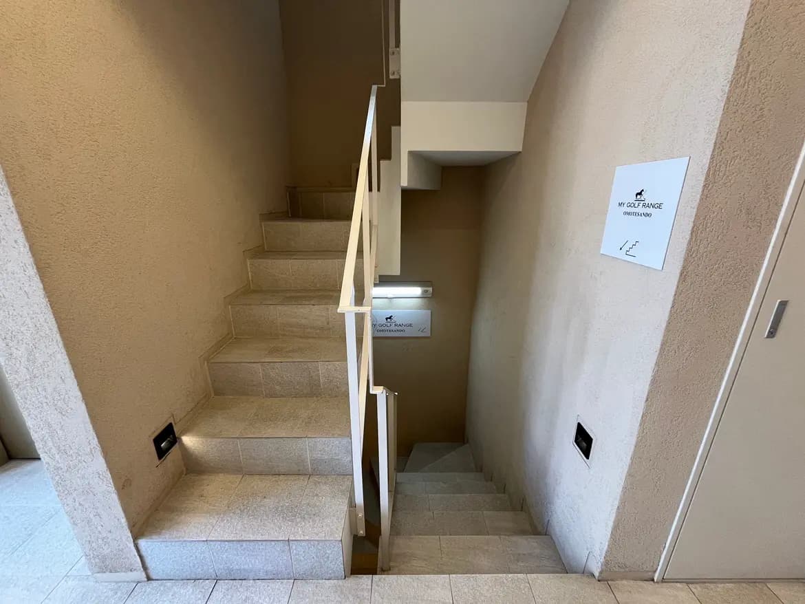 - 明るい色の壁に囲まれた階段、手すりがついている屋内の階段、ゴルフレンジへの案内 sign が掲示されている様子