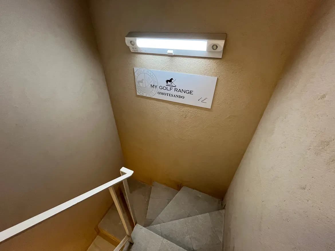 - 明るい照明の下、出入り口の看板「MY GOLF RANGE OMOTESANDO」が掲げられた階段の内部画像、淡い色合いの壁と手すりが特徴的