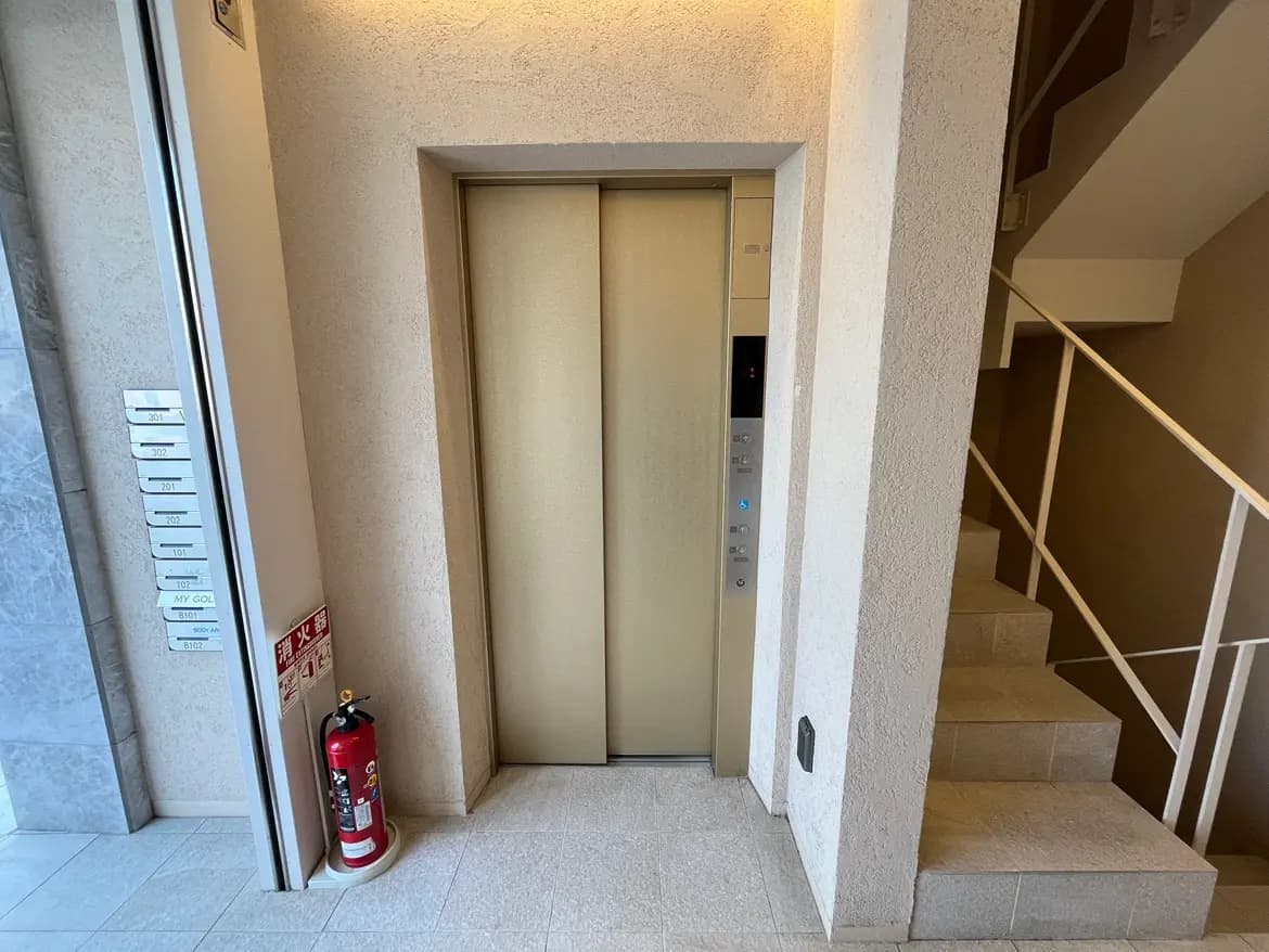- 建物内のエレベーターの入口、周囲には階段と消火器が配置されたシンプルなエントランスの風景