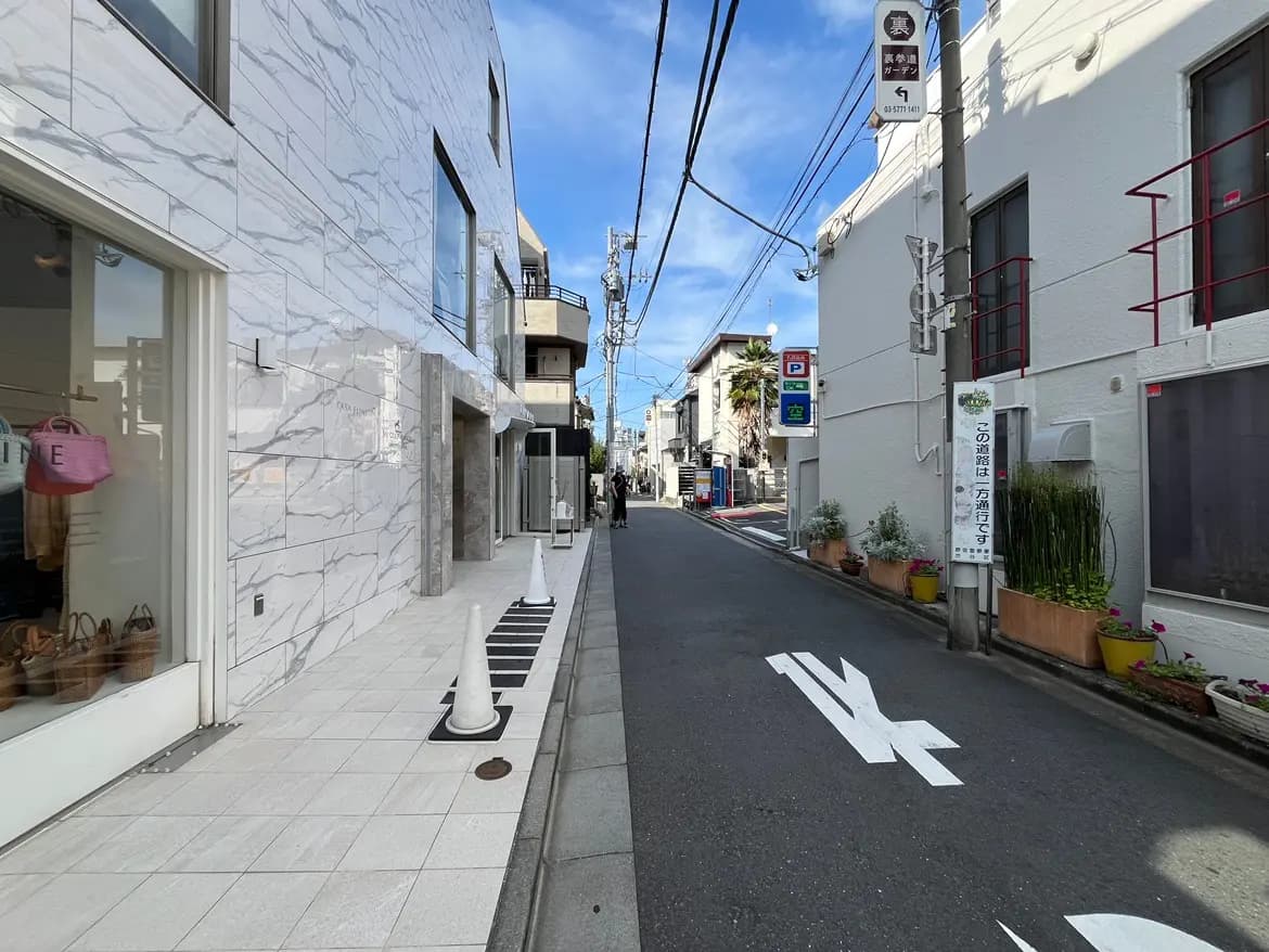- 大理石風の外壁を持つスタイリッシュな店舗入口、小道と白いコーン、大きな窓から見える店内の商品ディスプレイが特徴の都市風景
