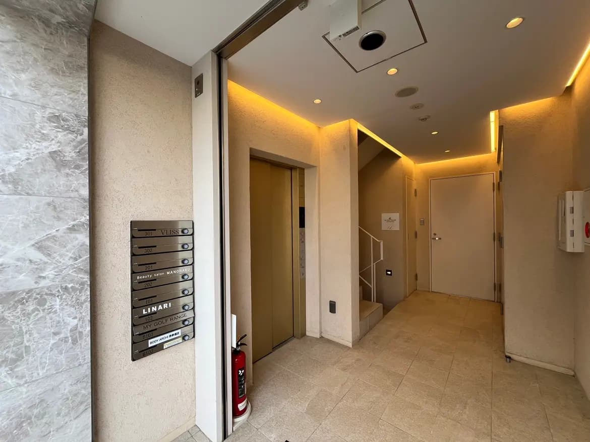 - モダンなエントランスホール、エレベーターの表示板、石材の壁と落ち着いた照明が特徴の空間