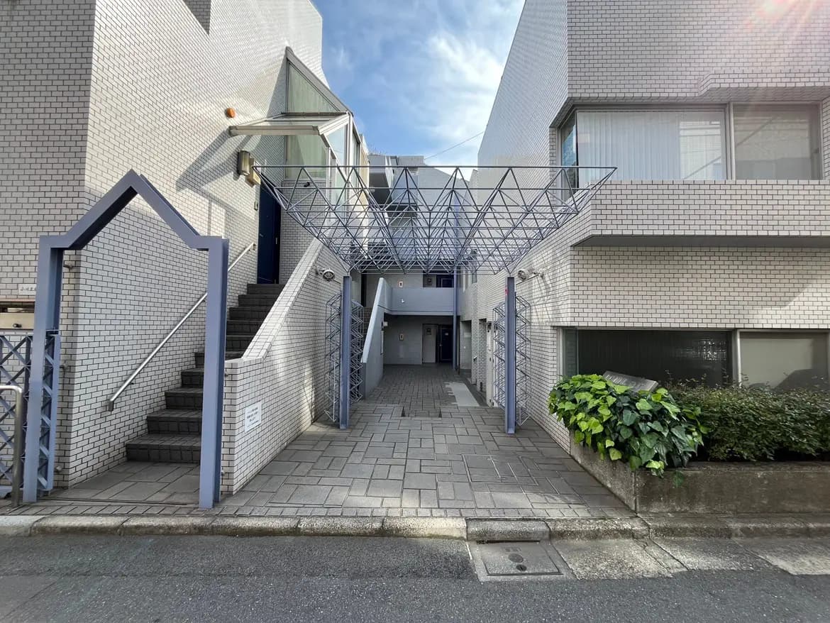 - 近代的なデザインのアパートエントランス、白いタイルの外壁と屋根付き階段、周囲に緑がある景観