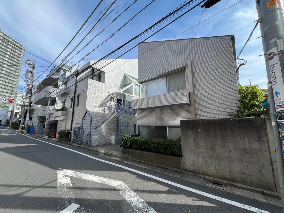 - モダンなデザインの建物外観、白いタイル張りの壁と大きな窓、周囲に街路と電線が走る都市風景