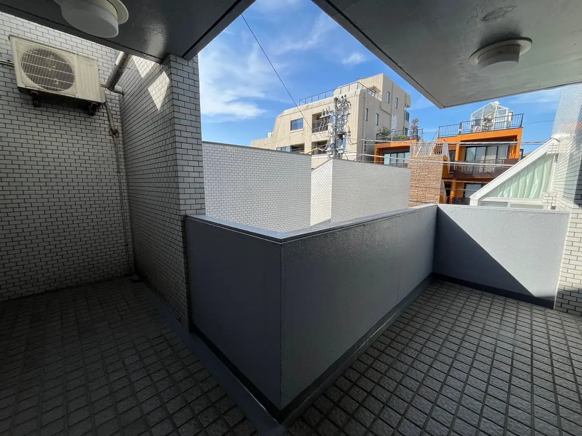 - 明るい空と隣接する建物を見渡せるバルコニー、タイル張りの床とコンクリートの壁が特徴の屋外スペース