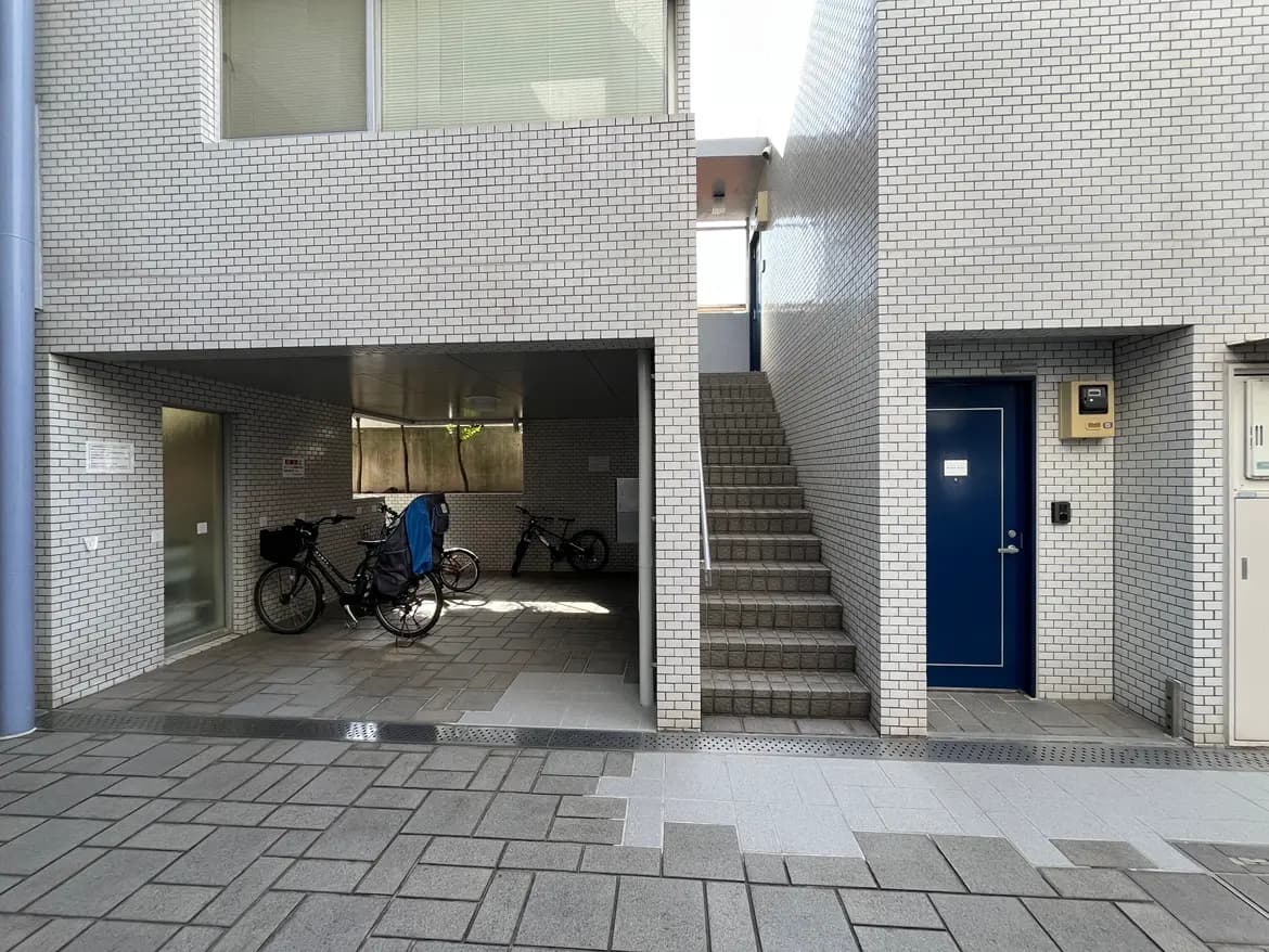 - シンプルでモダンな住宅の外観、駐輪スペースと階段、白いタイルの壁と青いドアがあるエントランス