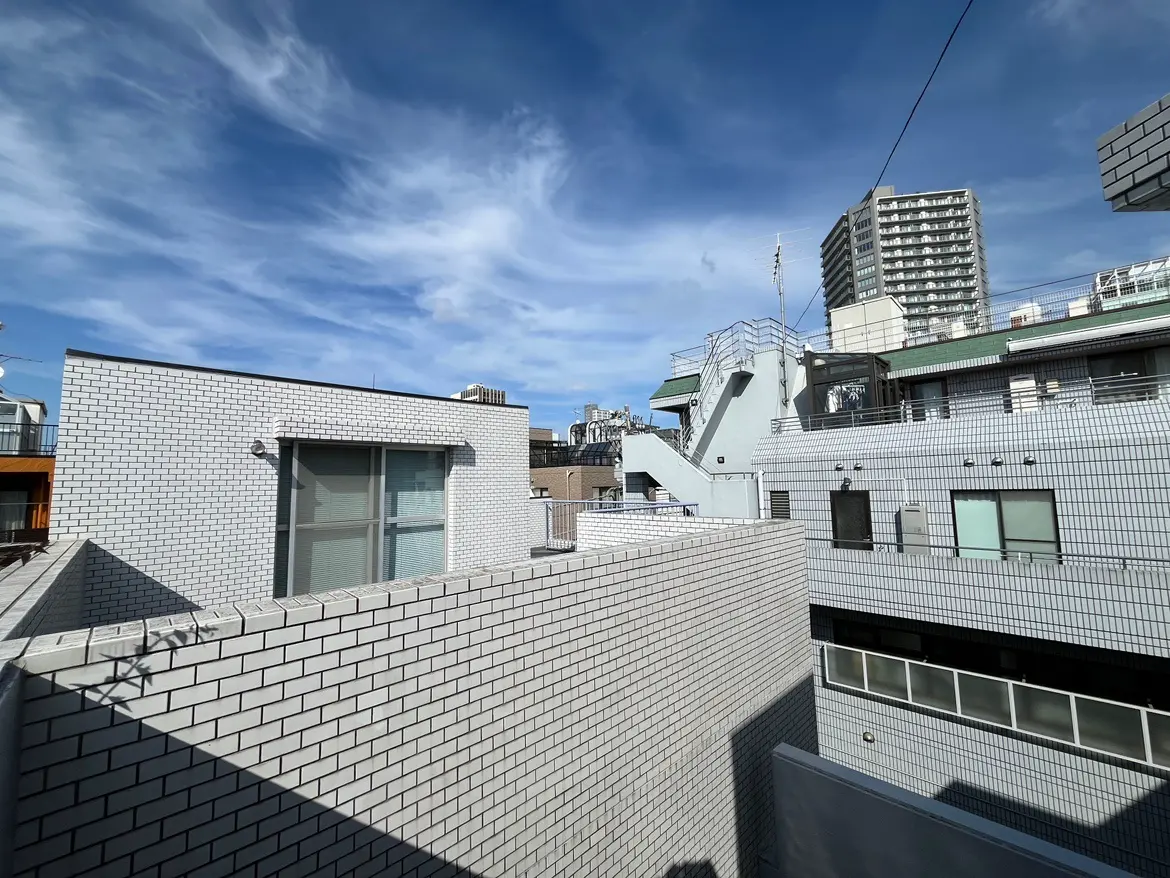 - 青空と白いレンガの建物、隣接する建物が見える都市の風景、陽射しが柔らかく当たる屋上の景観