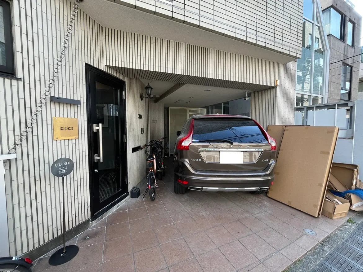 - モダンな建物の入り口、黒いドアと看板、駐車場に置かれた自転車と車、周囲に散らばる段ボール箱
