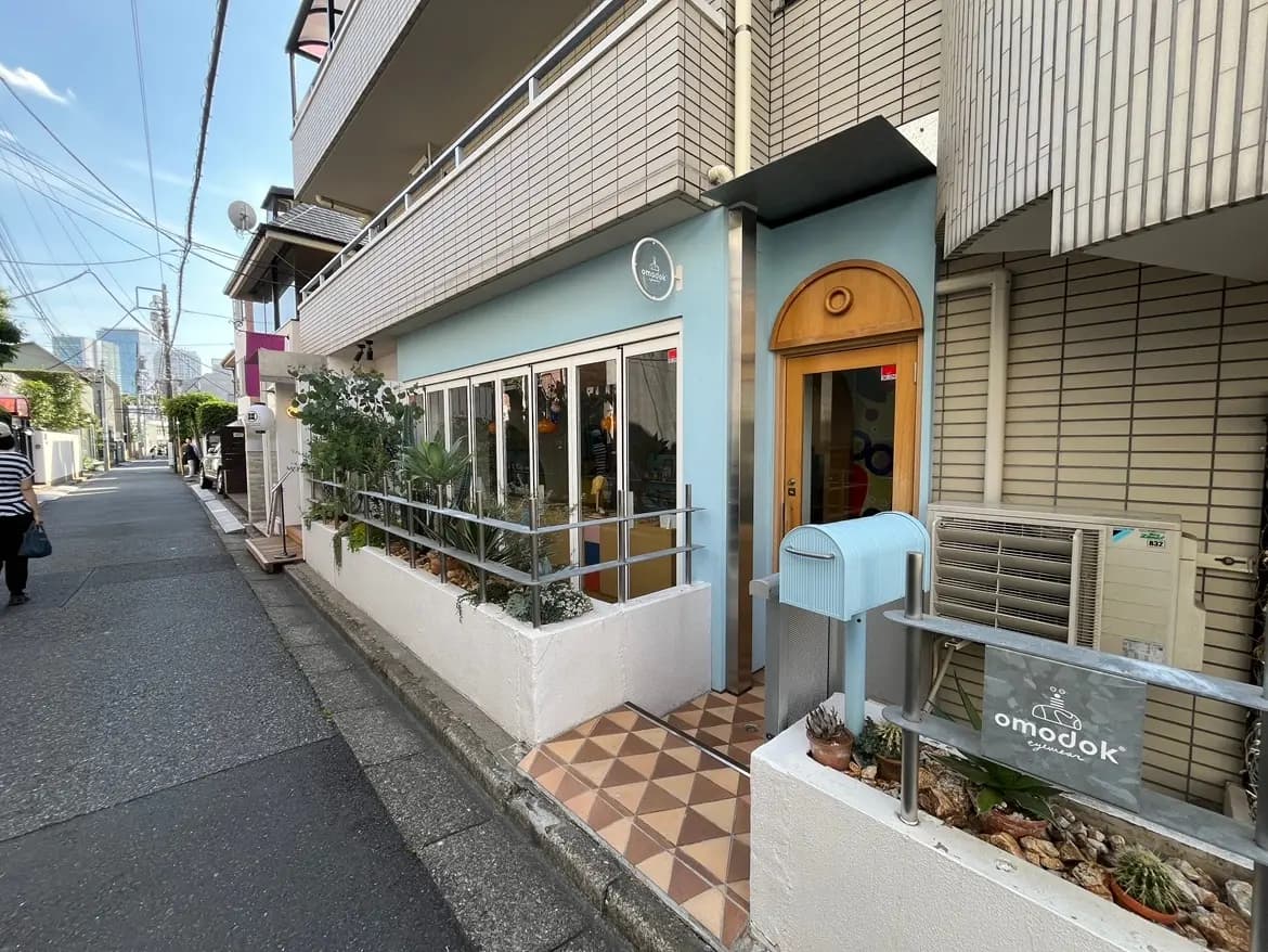 - 都会の路地に位置するカフェの外観、青い壁と木製のドア、植物が飾られた縁台やオシャレな郵便受けが特徴的なショップファサード