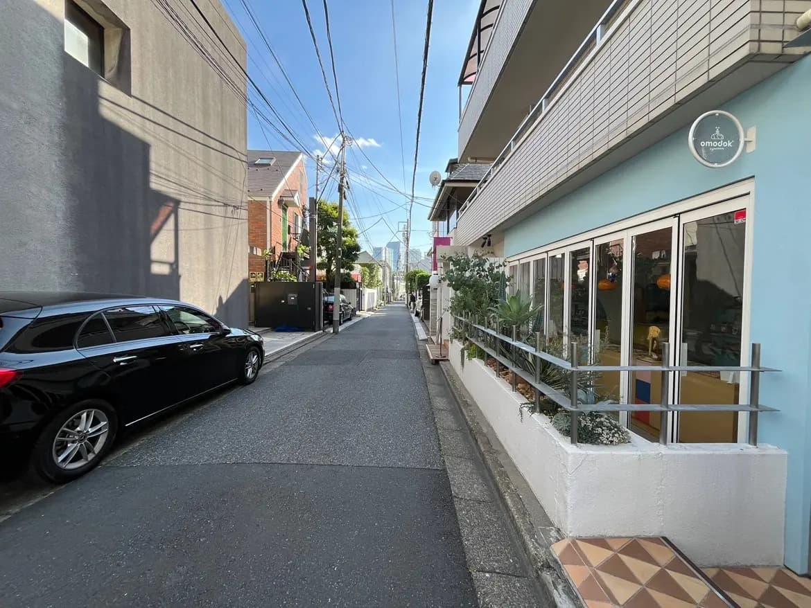 - 都市の路地に沿ったカフェの外観、植物が飾られたフェンスと開放的な窓、大きな空と電線が見える景色