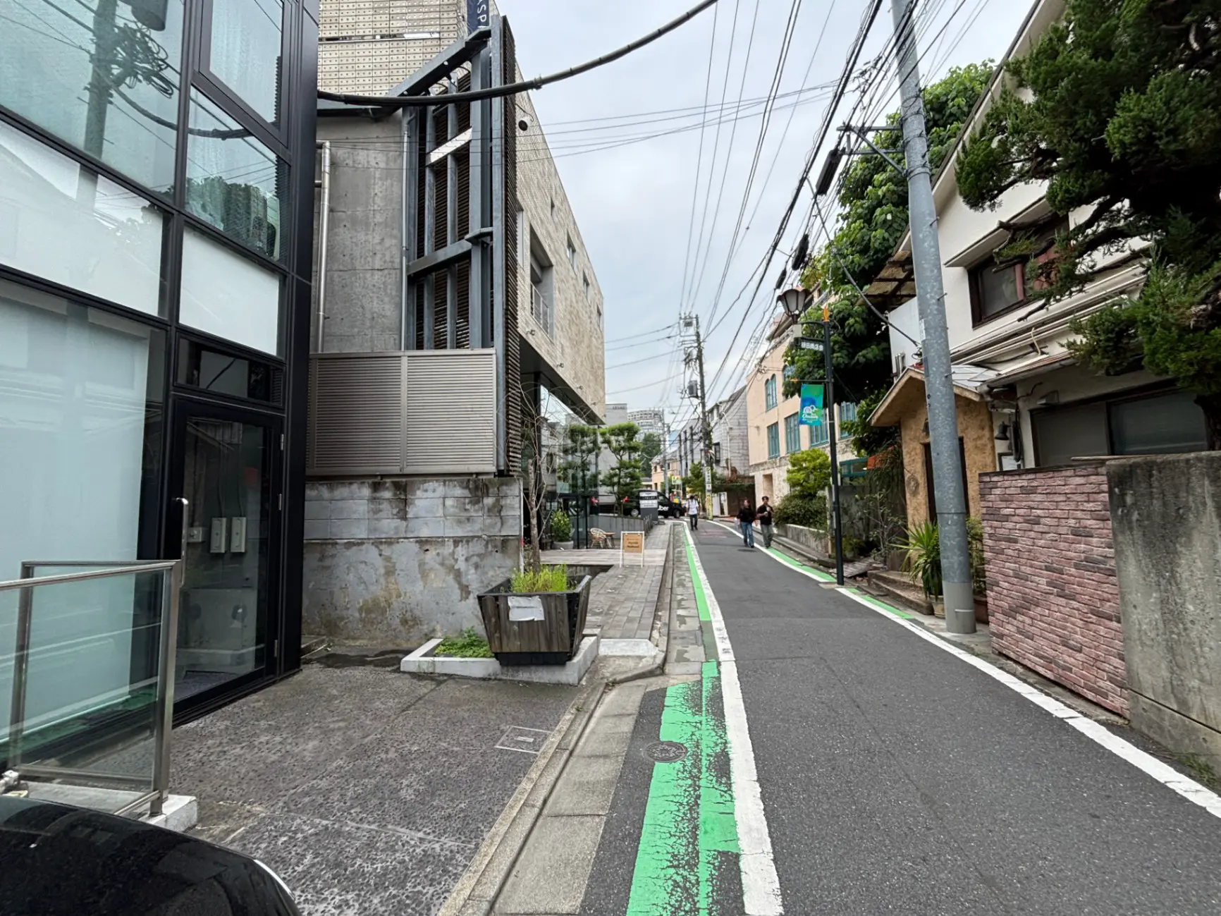 - 現代的なコンクリート建築とガラスのファサード、街並みが写る狭い道路と歩道、電柱と電線が交差する風景