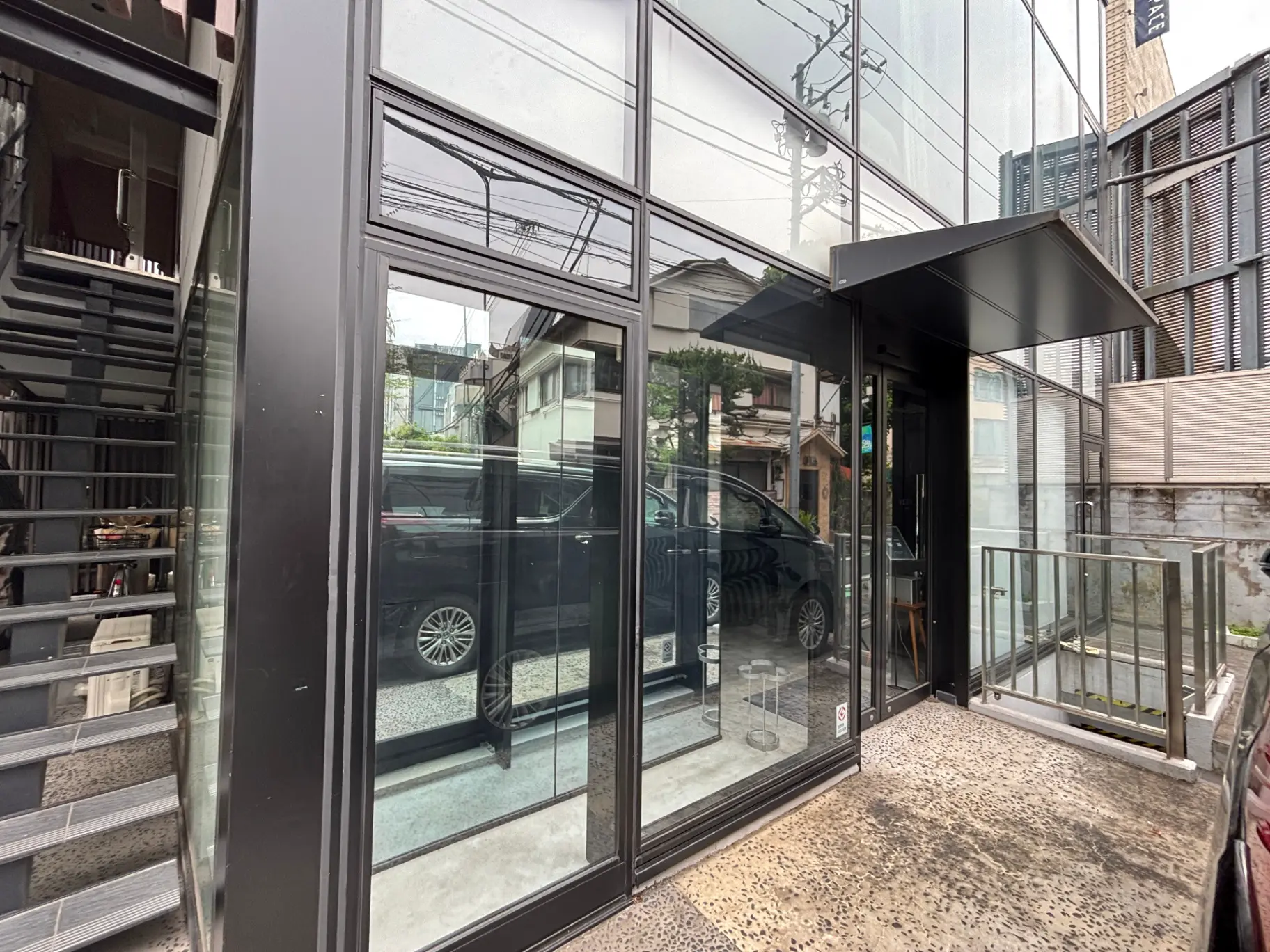 - ガラスの外観を持つモダンな建物のエントランス、隣に階段があり、周囲には車と建物がある景観