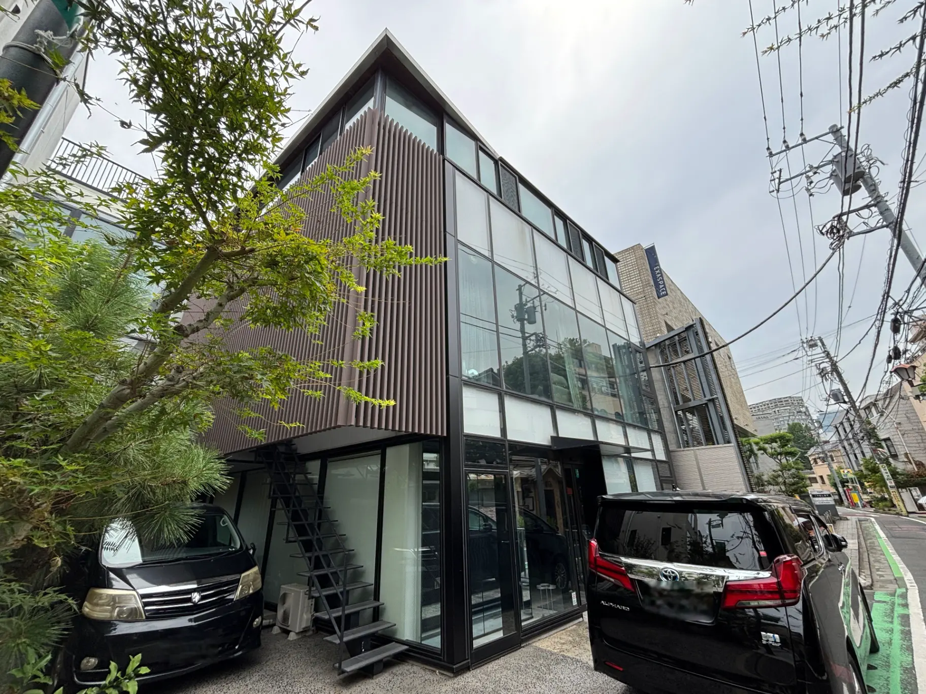 - 近代的なデザインの住宅外観、黒い車と植栽がある立地、ガラスのファサードと階段が特徴的な建物