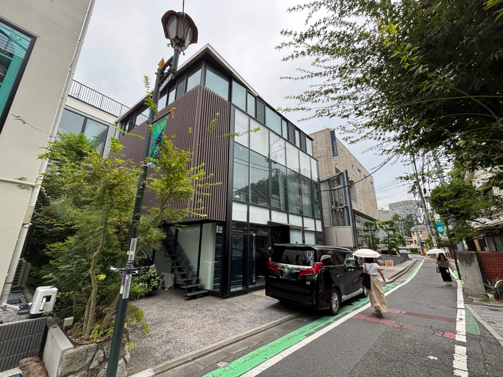 - モダンなデザインのビル、ガラスと木材の外観、周囲の緑と街灯、歩行者が行き交う道路の景観