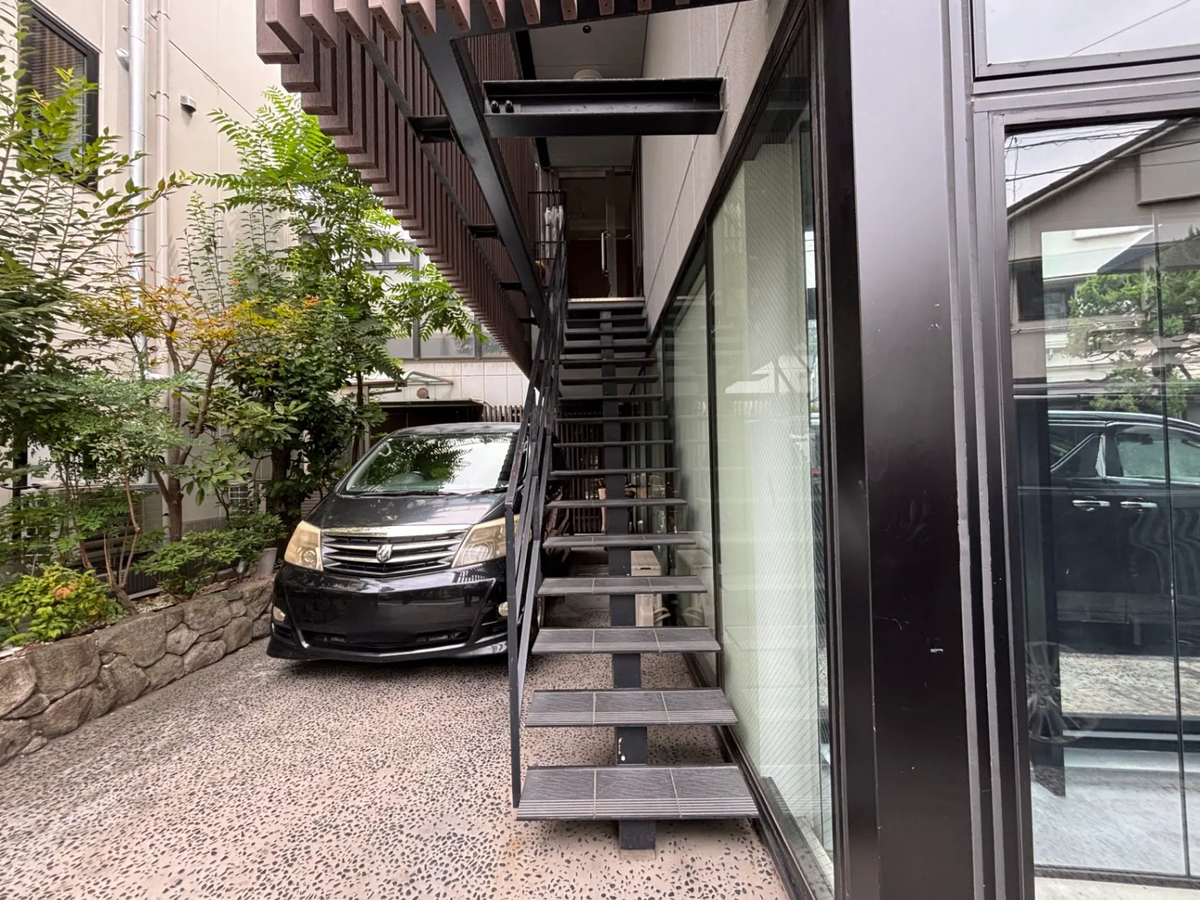 - 鉄製の階段が付いた現代的な住宅の外観、隣には黒いミニバンと緑豊かな植栽が見える様子
