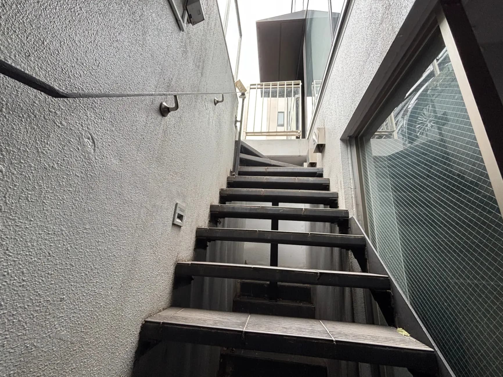 - コンクリートの壁に囲まれた木製の階段、上に向かって続く明るい手すりと隣接する窓が特徴的な風景