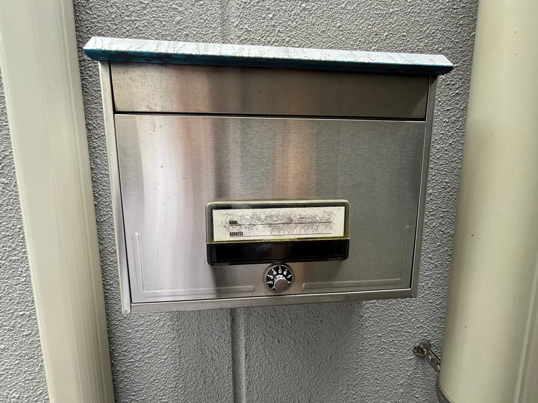 - ステンレス製の郵便受け、ダイヤルロック付き、灰色の壁面に取り付けられたシンプルでモダンなデザイン