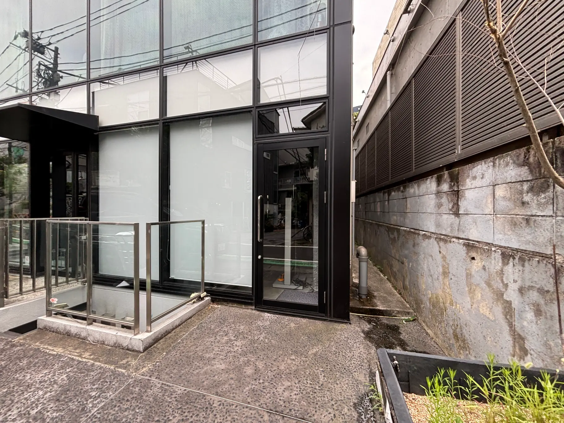 モダンなデザインの建物のエントランス、ガラスのファサードと金属製の手すり、周囲に自然な素材が調和する景観