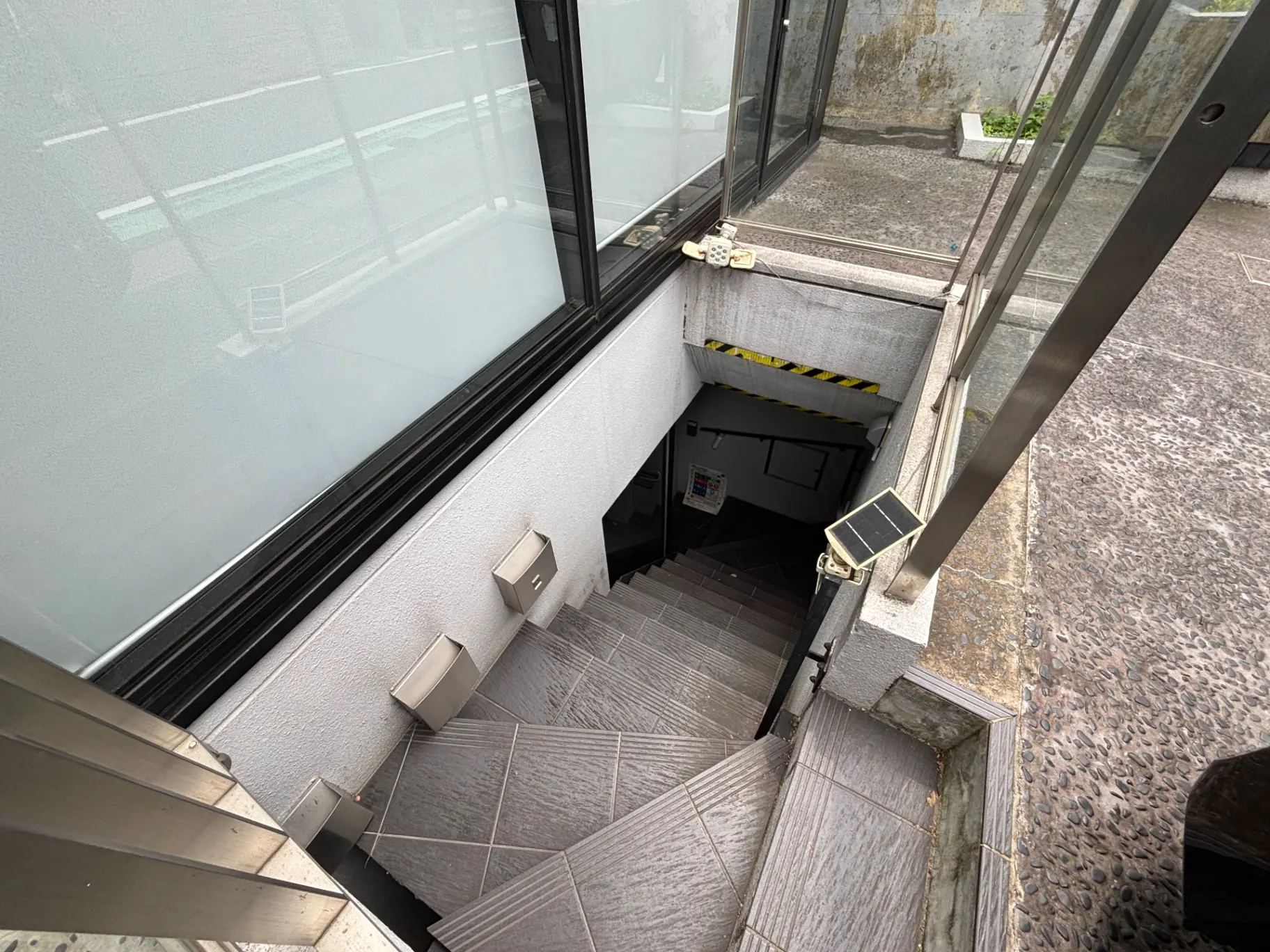 - 照明が設置された階段の上から見下ろした風景、白い壁とグレーのタイルの階段、下階へのアクセスを示す出入り口