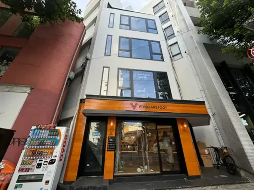 - シンプルでスタイリッシュな外観の店舗ビル、木目調の看板とガラス窓、周囲にある自販機と自転車のある風景