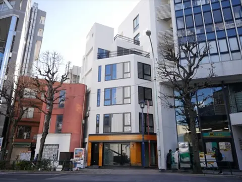 - 都会の街並みの中に佇むモダンなビル、白い外壁と大きな窓、隣接する店舗との対比が特徴的な外観