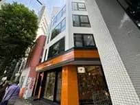 - 現代的なデザインのビル外観、オレンジ色の看板が目を引くストリートの風景、周囲に人々が行き交う様子