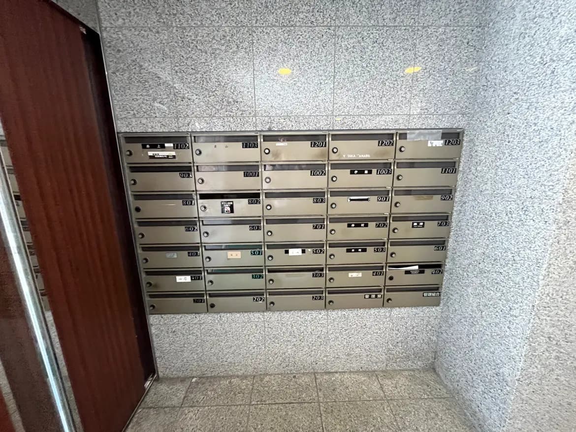 - マンションのエントランスにある郵便受け、整然と配置された金属製のボックス、周囲にはグレーのタイル壁と木製のドアが見える
