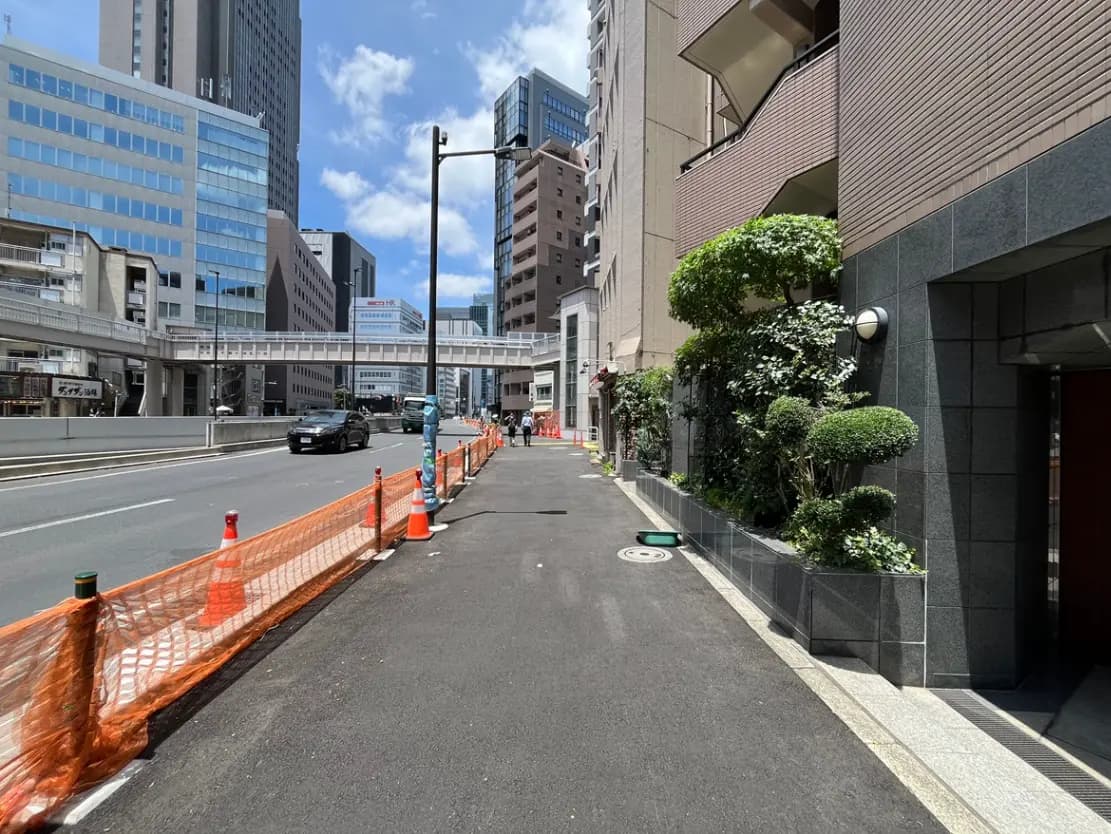 - 都市部の街並み、交通量のある道路と歩道、工事中のオレンジのバリケード、周囲の高層ビルと青空の景観