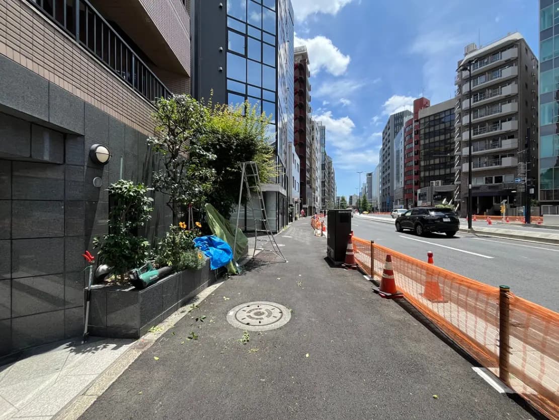 - 都市部の歩道沿いに並ぶ高層ビル群、緑の植栽と作業用道具が置かれた景観、青空と白い雲の下の街並み