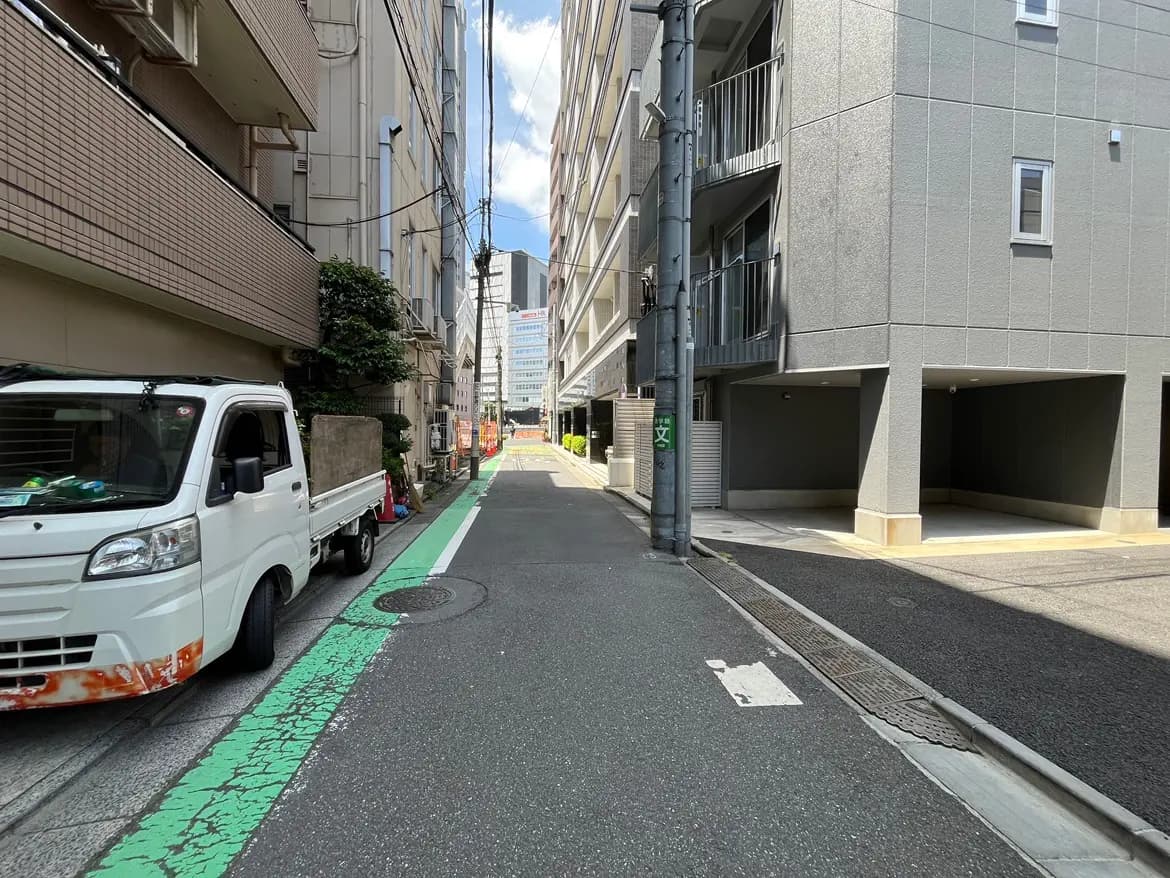 - 都市の狭い路地に停まっている白いトラックと、その奥に見えるビル群の風景、青空と白い雲が広がる昼間のシーン