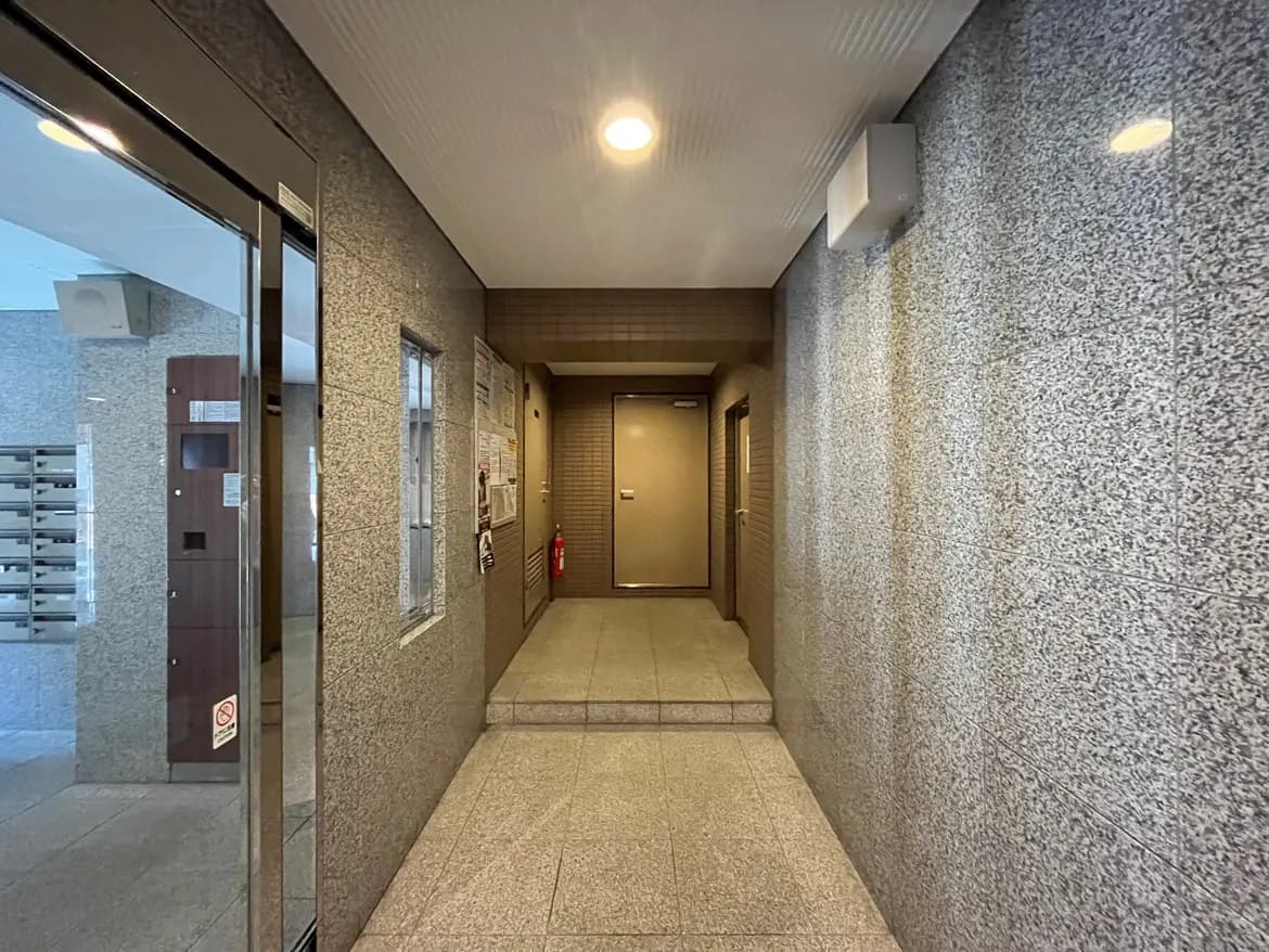 - モダンな建物のエントランス、グレーのタイルの壁と床、明るい照明、郵便受けや掲示板がある共用スペース