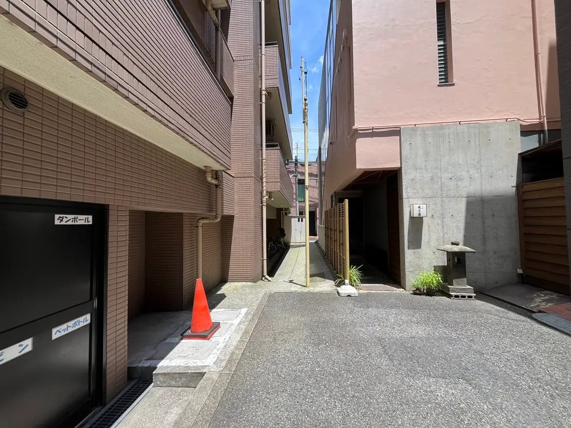 - 住宅街の狭い路地、左側にあるダンボール置き場とペットボトル回収ボックス、右側にはコンクリート製の壁と竹のフェンスが見える風景