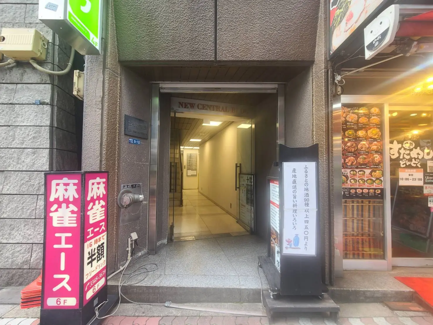 - 入口に「NEW CENTRAL BUILDING」の看板が掲げられたビルの外観、麻雀店の看板と飲食店のメニューが並ぶ街の風景