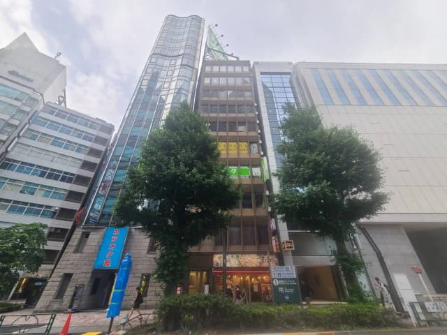 - 都市の中心に位置するビル群、カラオケ店の外装や飲食店の看板が見える風景、周囲に緑の樹木が並ぶ街並み