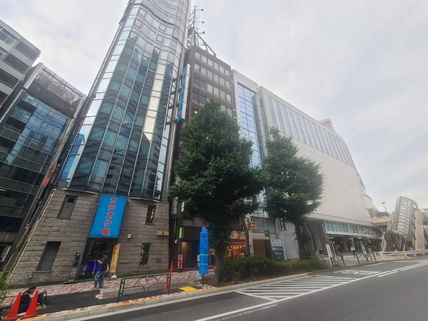 - カラオケ館の看板が見えるビルと周囲の商業施設、道路には歩行者と植栽が配置された街並みの風景
