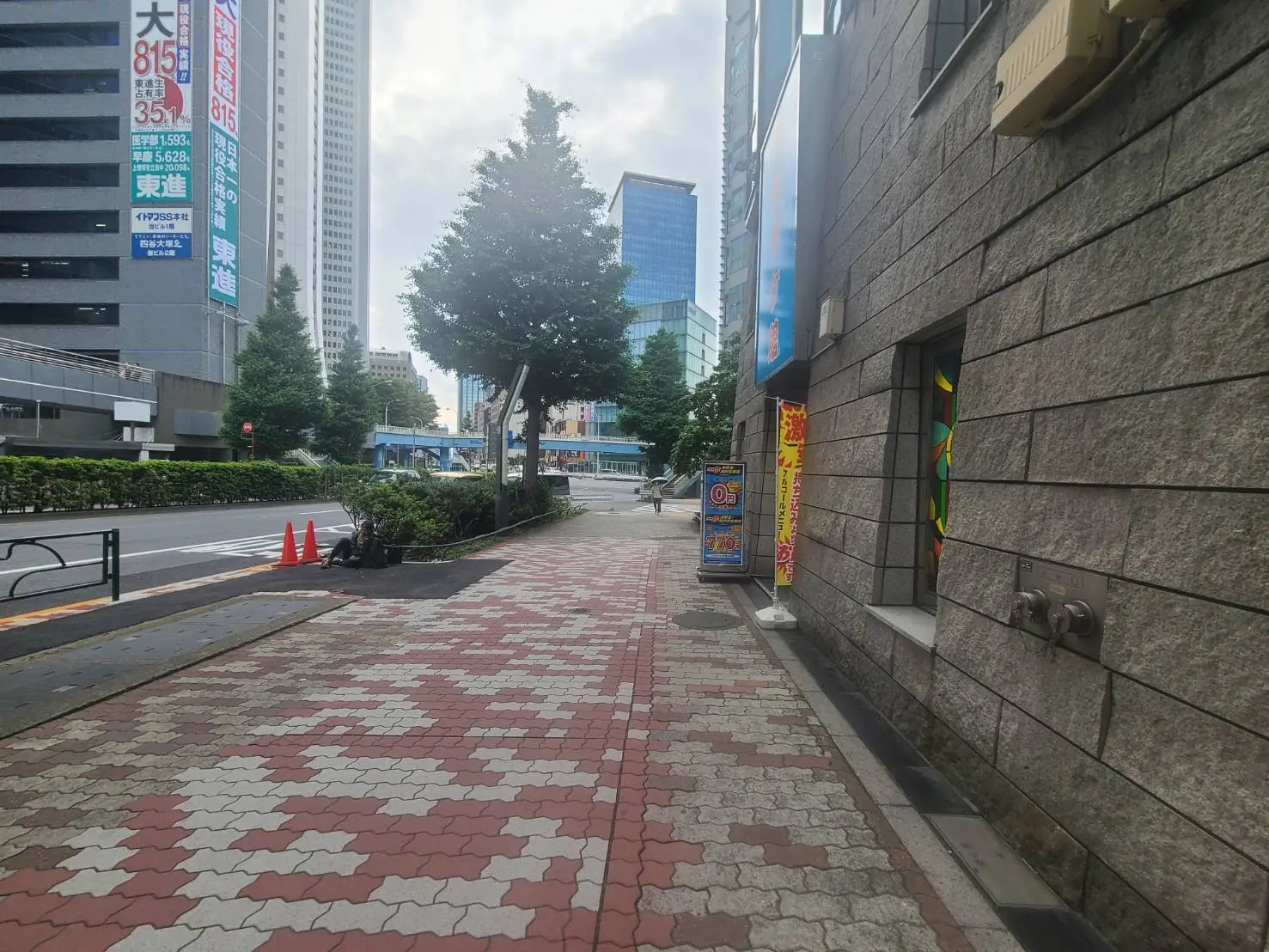 - 都市の歩道、タイルの舗装、ビルの隙間にある樹木、交通表示と広告が見える風景
