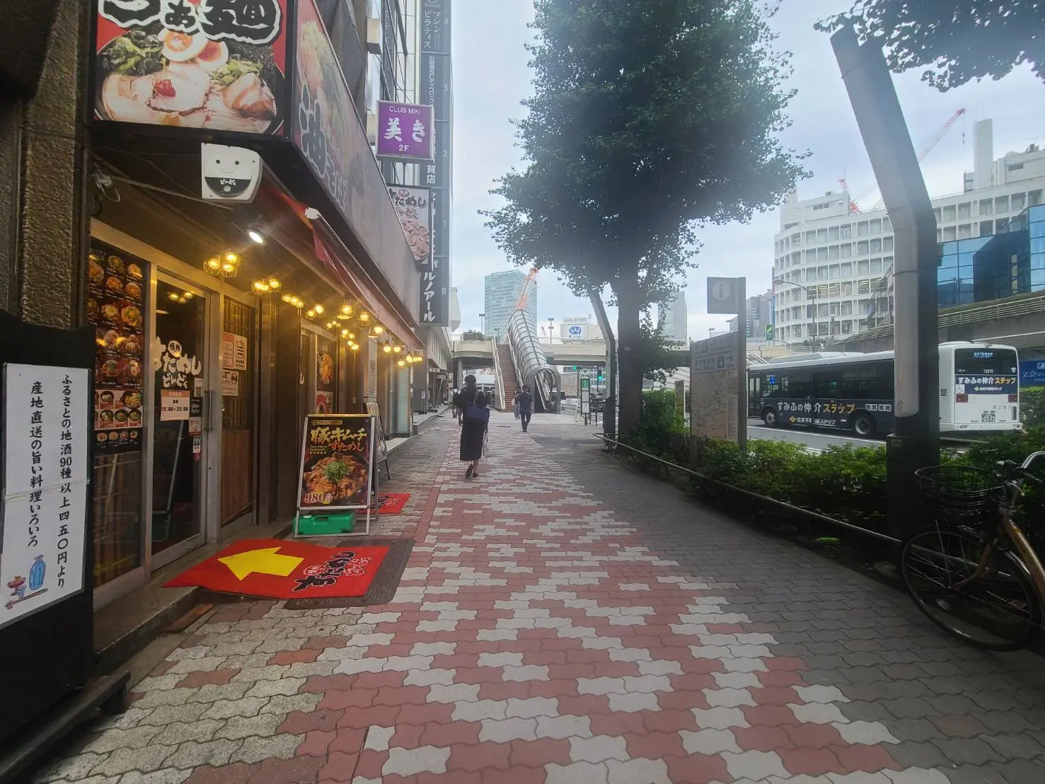 - 都市の通り沿いにある飲食店の外観、立て看板と照明、歩道には人々と緑の木々が並ぶ風景