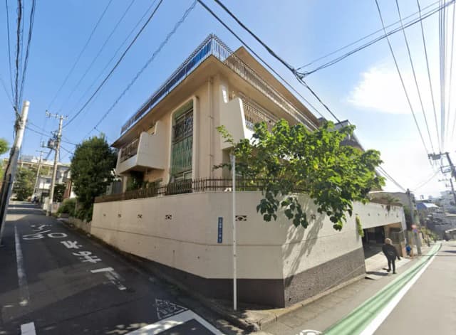 - 住宅街に位置する角地の建物、緑豊かな木々と電線の見える青空の下の外観、通行人が歩く街並み