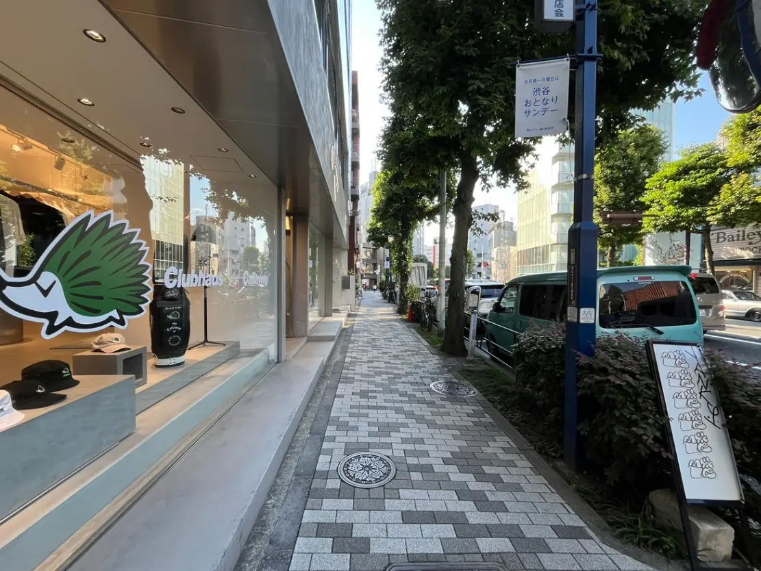 - 都会の商業エリアにある「Clubhaus」の店舗外観、周囲の緑と繁忙な歩道が印象的な風景