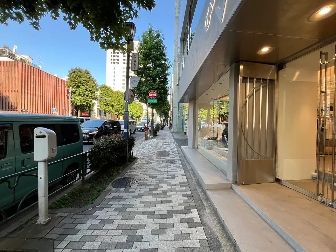 - 街並みの歩道とショップの前景、青空の下で車や緑の植栽が見える風景
