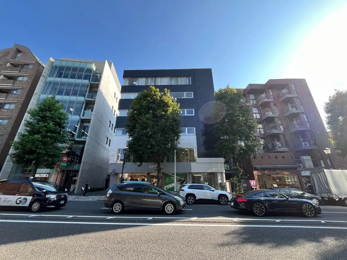 - 現代的なビルが並ぶ都市の風景、左側にはガラス張りの建物と緑の木、右側には複数のバルコニーがあるレンガ造りの建物と車が停まっている様子
