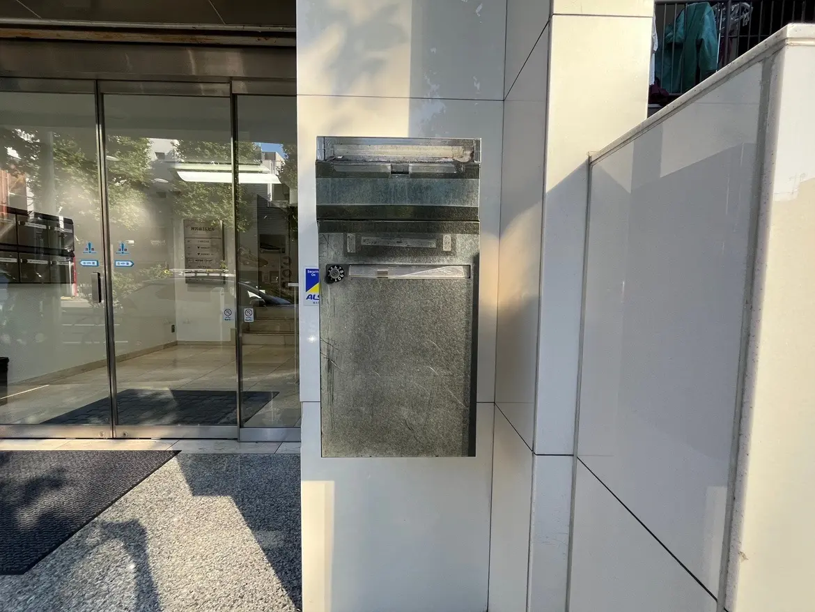 - モダンな建物の入口に設置されたシンプルなデザインの郵便受け、ガラス扉越しに見える明るいロビーの様子