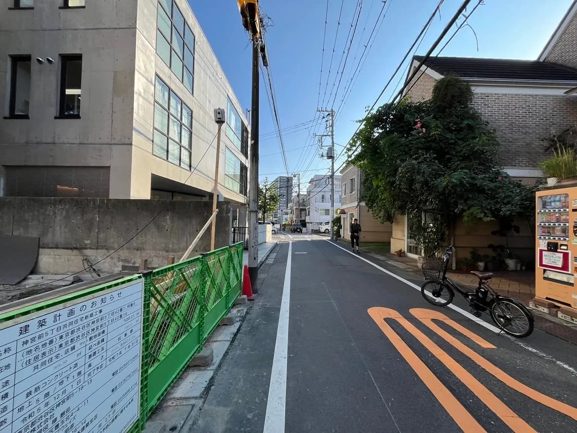 - 緑のフェンスが設置された工事現場と近隣の住宅、左側に自転車が停められた静かな街道と青空が広がる風景